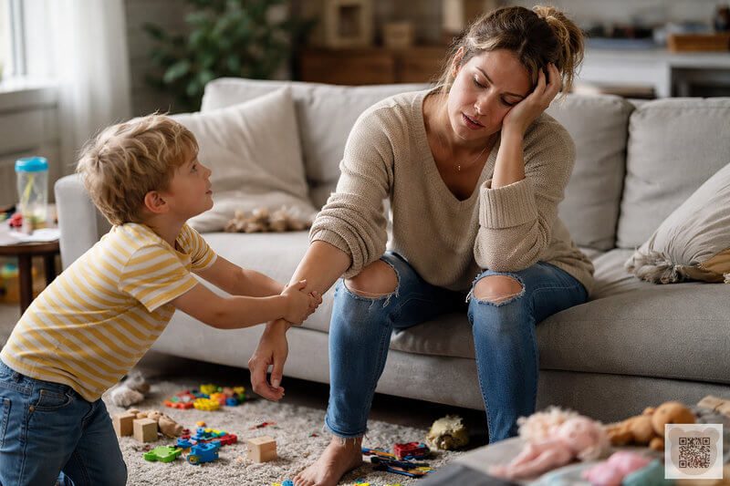 Exhausted mother sitting on a sofa while her toddler pulls her arm because the child won't play alone; featuring expert teacher advice and tips for encouraging independent play.